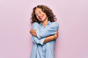 Middle age beautiful woman wearing casual denim shirt standing over pink background Hugging oneself happy and positive, smiling confident. Self love and self care - obrazy, fototapety, plakaty