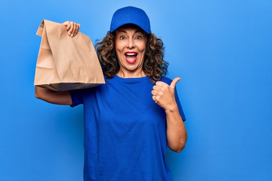 Middle age beautiful delivery woman holding deliver paper bag with takeaway food pointing thumb up to the side smiling happy with open mouth