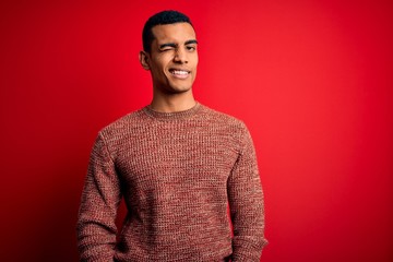 Young handsome african american man wearing casual sweater standing over red background winking...