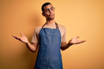 Young handsome african american shopkeeper man wearing apron over yellow background clueless and...