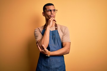 Young handsome african american shopkeeper man wearing apron over yellow background with hand on chin thinking about question, pensive expression. Smiling with thoughtful face. Doubt concept. - obrazy, fototapety, plakaty
