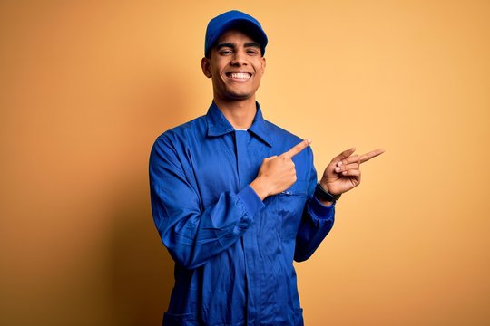 Young african american mechanic man wearing blue uniform and cap over yellow background smiling and looking at the camera pointing with two hands and fingers to the side.