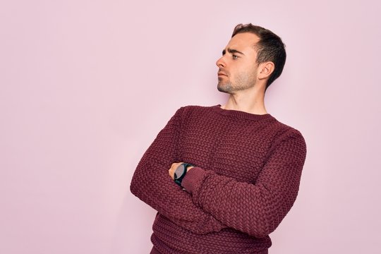 Young handsome man with blue eyes wearing casual sweater standing over pink background looking to the side with arms crossed convinced and confident