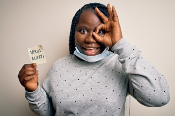 African american woman wearing medical mask holding reminder with virus alert message with happy face smiling doing ok sign with hand on eye looking through fingers