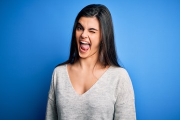 Fototapeta premium Young beautiful brunette woman wearing casual sweater standing over blue background winking looking at the camera with sexy expression, cheerful and happy face.