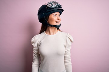 Young beautiful motorcyclist woman with blue eyes wearing moto helmet over pink background looking away to side with smile on face, natural expression. Laughing confident.