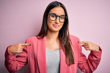 Young beautiful brunette businesswoman wearing jacket and glasses over pink background looking...