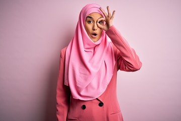 Young beautiful brunette businesswoman wearing pink muslim hijab and business jacket doing ok gesture shocked with surprised face, eye looking through fingers. Unbelieving expression.