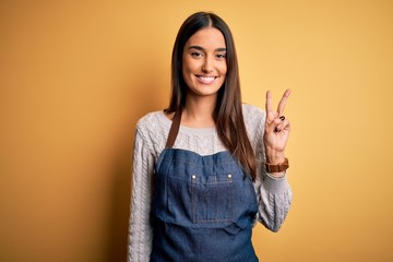 Young beautiful baker woman wearing apron uniform cooking over yellow background smiling with happy face winking at the camera doing victory sign. Number two.