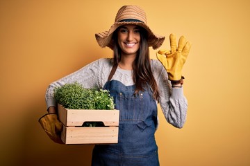 Young beautiful brunette gardener woman wearing apron and hat holding box with plants doing ok sign...