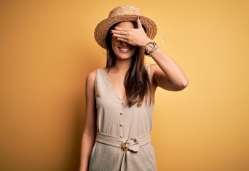 Young beautiful brunette woman on vacation wearing casual dress and hat smiling and laughing with hand on face covering eyes for surprise. Blind concept.