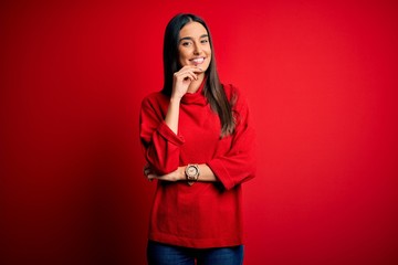 Young beautiful brunette woman wearing casual sweater over isolated red background looking confident at the camera smiling with crossed arms and hand raised on chin. Thinking positive.