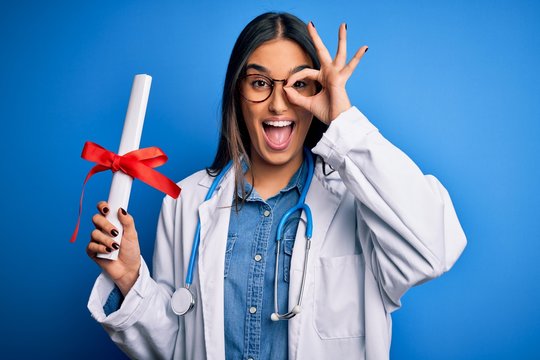Young beautiful brunette doctor woman wearing glasses and coat holding diploma degree with happy face smiling doing ok sign with hand on eye looking through fingers