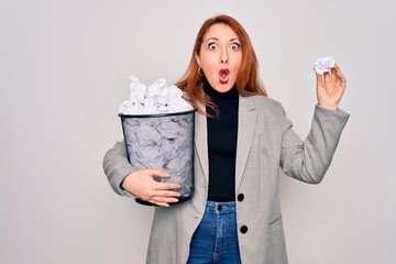 Young beautiful redhead businesswoman holding full paper bin with crumpled papers scared in shock with a surprise face, afraid and excited with fear expression