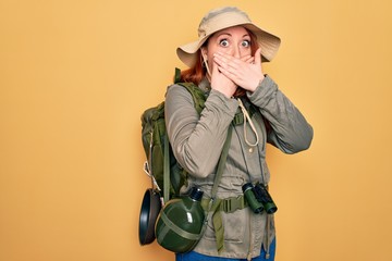 Young redhead backpacker woman hiking wearing backpack and hat over yellow background shocked covering mouth with hands for mistake. Secret concept.