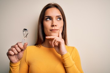 Young beautiful brunette woman holding eyelash curler over isolated white background serious face thinking about question, very confused idea