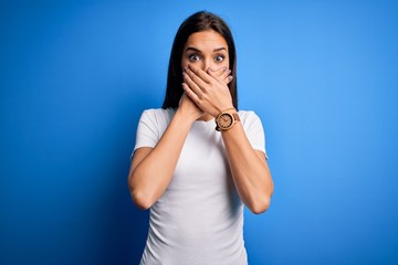 Young beautiful brunette woman wearing white casual t-shirt standing over blue background shocked covering mouth with hands for mistake. Secret concept.