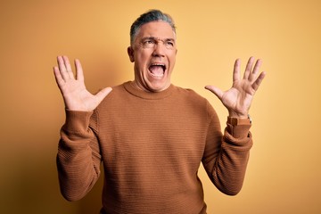 Middle age handsome grey-haired man wearing casual sweater over yellow background crazy and mad shouting and yelling with aggressive expression and arms raised. Frustration concept.