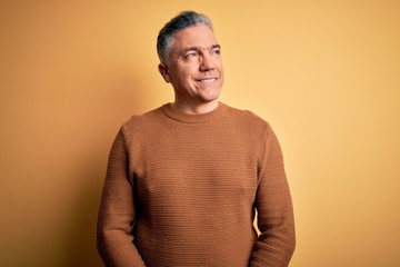 Obraz premium Middle age handsome grey-haired man wearing casual sweater over yellow background looking away to side with smile on face, natural expression. Laughing confident.