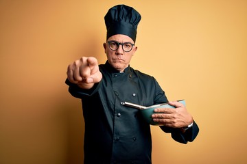 Middle age handsome grey-haired chef man wearing cooker uniform and hat using whisk pointing with finger to the camera and to you, hand sign, positive and confident gesture from the front