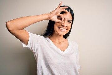 Young beautiful hispanic woman wearing casual white t-shirt over isolated background doing ok gesture with hand smiling, eye looking through fingers with happy face.