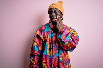Obraz premium Young handsome african american man wearing colorful coat and cap over pink background touching mouth with hand with painful expression because of toothache or dental illness on teeth. Dentist