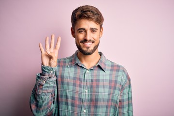 Young handsome man with beard wearing casual shirt standing over pink background showing and pointing up with fingers number four while smiling confident and happy.