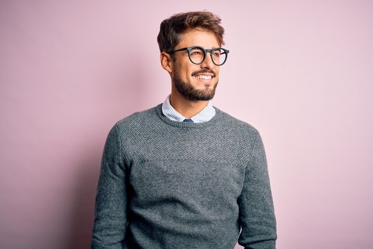 Young handsome man with beard wearing glasses and sweater standing over pink background looking away to side with smile on face, natural expression. Laughing confident.