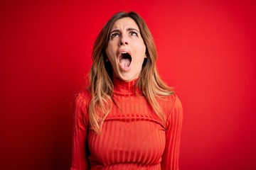 Young beautiful brunette woman wearing casual turtleneck sweater over red background angry and mad screaming frustrated and furious, shouting with anger. Rage and aggressive concept.