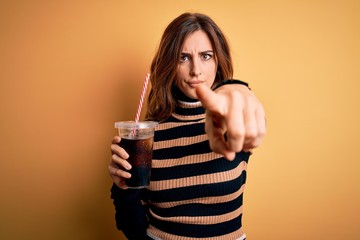 Young beautiful brunette woman drinking glass with cola refreshment using straw pointing with finger to the camera and to you, hand sign, positive and confident gesture from the front