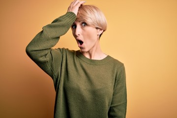 Young blonde woman with modern short hair wearing casual sweater over yellow background surprised...