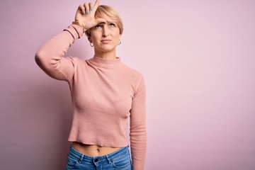 Young blonde woman with short hair wearing casual turtleneck sweater over pink background making fun of people with fingers on forehead doing loser gesture mocking and insulting.