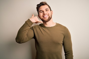 Young blond man with beard and blue eyes wearing green sweater over white background smiling doing phone gesture with hand and fingers like talking on the telephone. Communicating concepts.