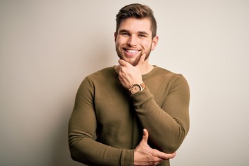 Young blond man with beard and blue eyes wearing green sweater over white background looking confident at the camera smiling with crossed arms and hand raised on chin. Thinking positive.