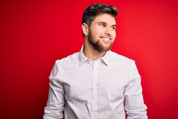 Obraz premium Young blond businessman with beard and blue eyes wearing elegant shirt over red background looking away to side with smile on face, natural expression. Laughing confident.