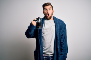 Young blond man with beard and blue eyes wearing pajama making coffe using coffeemaker scared in shock with a surprise face, afraid and excited with fear expression