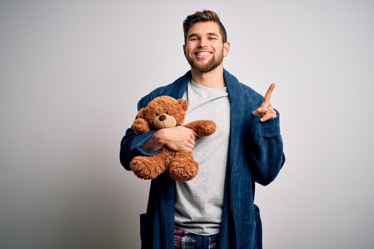 Young blond man with beard and blue eyes wearing pajama holding teddy bear surprised with an idea or question pointing finger with happy face, number one