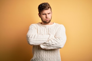 Young blond man with beard and blue eyes wearing white sweater over yellow background skeptic and...