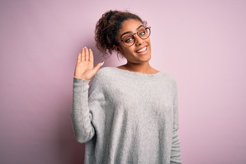Young beautiful african american girl wearing sweater and glasses over pink background Waiving saying hello happy and smiling, friendly welcome gesture