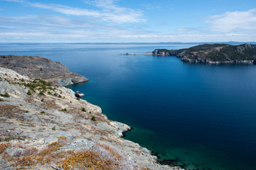 A large cove with deep blue ocean and sky. There are white fluffy clouds near the horizon, The cliffs are ragged and jagged rock. In the distance is a mountain covered in trees and a long rockface. 