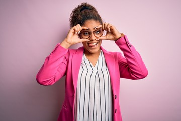 Beautiful african american businesswoman wearing jacket and glasses over pink background doing ok gesture like binoculars sticking tongue out, eyes looking through fingers. Crazy expression.