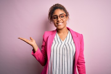 Beautiful african american businesswoman wearing jacket and glasses over pink background smiling cheerful presenting and pointing with palm of hand looking at the camera.