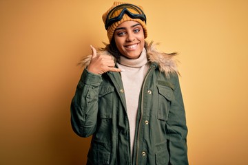 Young african american afro skier girl wearing snow sportswear and ski goggles smiling doing phone gesture with hand and fingers like talking on the telephone. Communicating concepts.