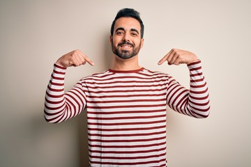 Young handsome man with beard wearing casual striped t-shirt standing over white background looking confident with smile on face, pointing oneself with fingers proud and happy.