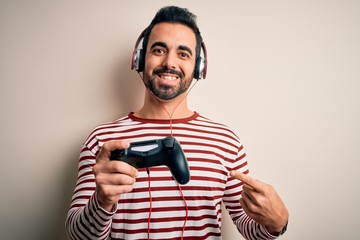 Young handsome gamer man with beard playing video game using joystick and headphones very happy pointing with hand and finger