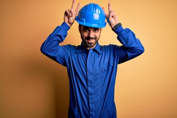 Mechanic man with beard wearing blue uniform and safety helmet over yellow background Posing funny...
