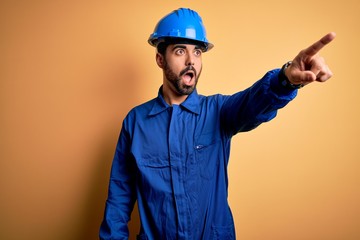 Mechanic man with beard wearing blue uniform and safety helmet over yellow background Pointing with finger surprised ahead, open mouth amazed expression, something on the front