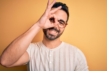 Young handsome man with beard wearing casual t-shirt and glasses over yellow background doing ok gesture with hand smiling, eye looking through fingers with happy face.