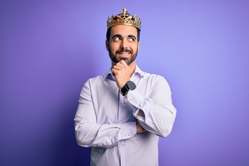 Young handsome man with beard wearing golden crown of king over purple background with hand on chin thinking about question, pensive expression. Smiling with thoughtful face. Doubt concept.