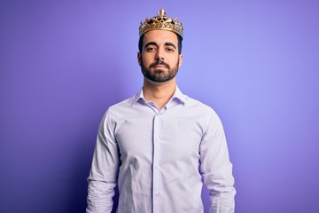Obraz premium Young handsome man with beard wearing golden crown of king over purple background Relaxed with serious expression on face. Simple and natural looking at the camera.